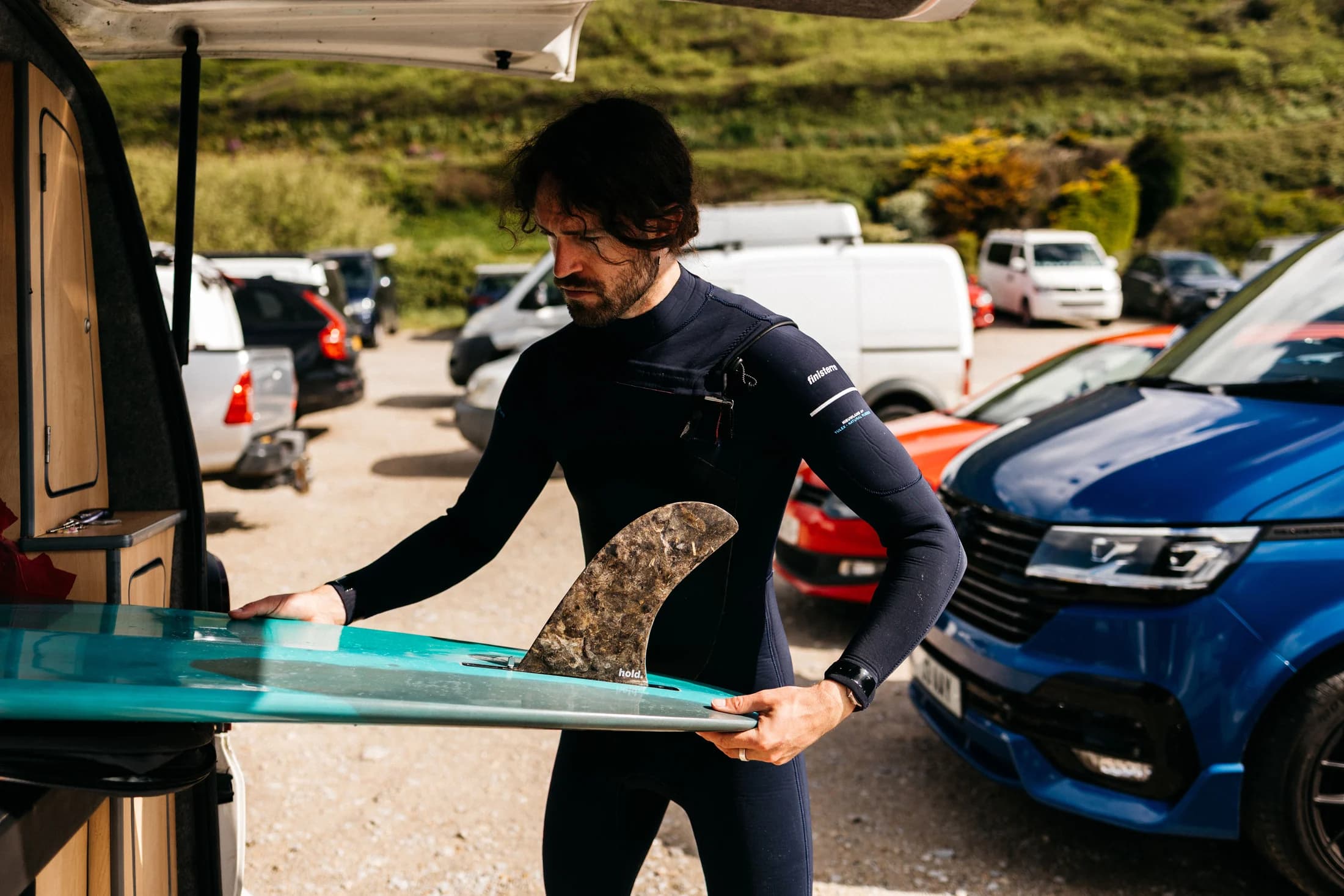 A surfer fitting a Hold fin beside a van before surfing.